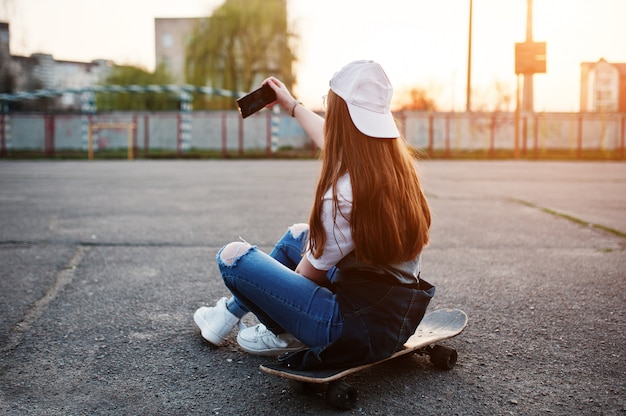 Joven adolescente urbana con patineta, usar anteojos, gorra y jeans rotos en el campo de deportes del patio en la puesta de sol haciendo selfie en el teléfono.