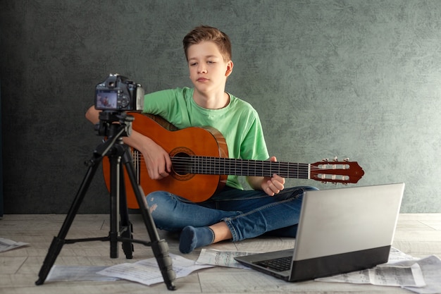 Joven adolescente videobloger aprende a tocar la guitarra en línea en la sala de estar, descansando en casa.