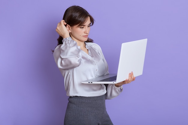 Joven adorable concentrada usando laptop contra la pared lila, estando ocupada, mira la pantalla de la computadora portátil, tocando su cabello mientras piensa en su trabajo, empresaria pensativa trabajando.