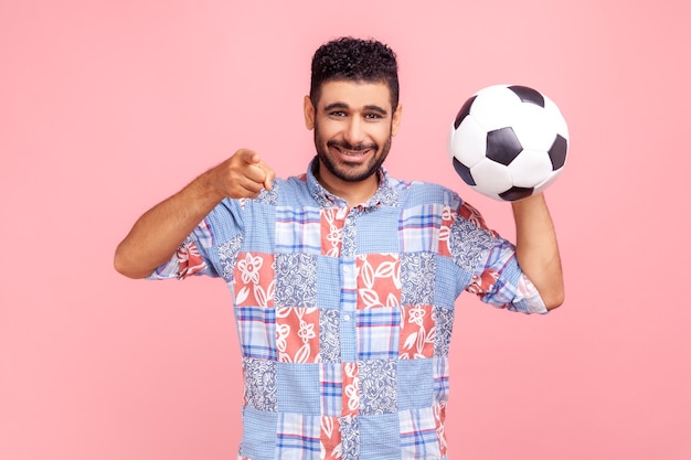 Un joven adulto con barba y una sonrisa dentuda con un pantalones azul de estilo casual apuntándote, sosteniendo una pelota de fútbol, mirando la cámara con una mirada feliz. Disparo de estudio interior aislado sobre fondo rosa.
