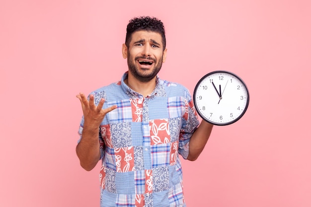 Un joven adulto disgustado, nervioso, barbudo, con el pelo oscuro gritando, mostrando el reloj de pared en la pantalla, fecha límite, usando una camisa azul de estilo informal. Disparo de estudio interior aislado sobre fondo rosa.