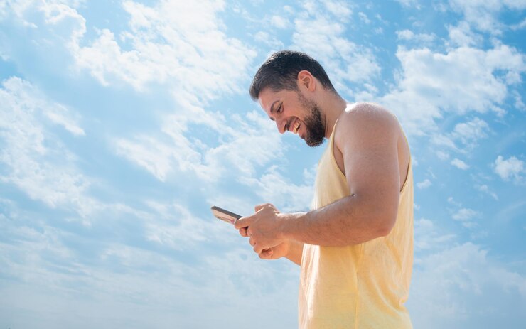 Joven adulto riendo en la playa enviando mensajes de texto con teléfono móvil y un bonito cielo azul soleado