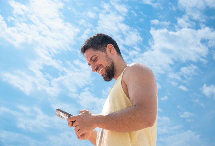 Un joven adulto sonriente en la playa con mensajes de texto en el teléfono móvil y un bonito cielo azul soleado