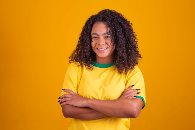Joven aficionado al fútbol brasileño negro con los brazos cruzados sobre el cuerpo Hermosa animadora brasileña con los brazos cruzados sobre fondo amarillo