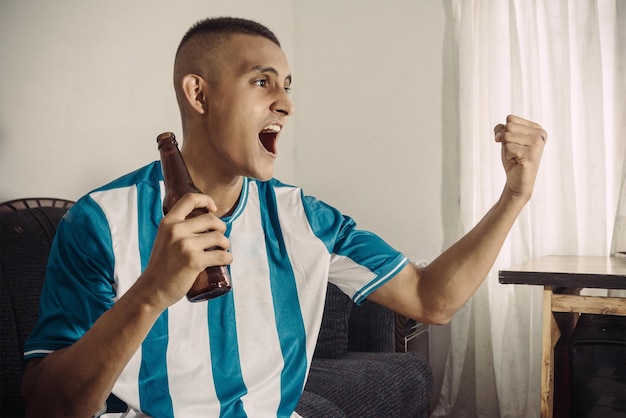 Joven aficionado al fútbol latinoamericano Celebra el gol Hombre Sostenga una cerveza Mire un televisor