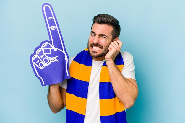 Joven aficionado a los deportes aislado de fondo azul cubriendo las orejas con las manos