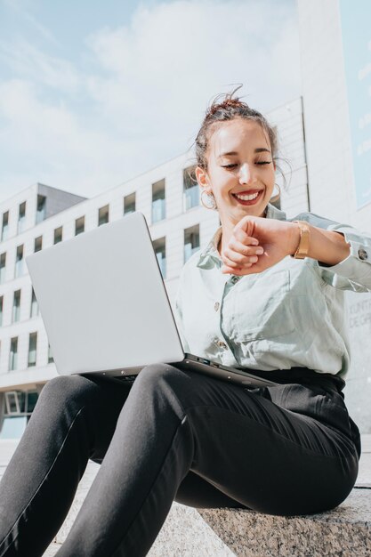 Joven africana árabe poc mujer de negocios comprobando su reloj mientras trabaja en su computadora portátil al aire libre en la ciudad, fuera de la oficina. Sonriendo feliz en el lugar de trabajo. Traje elegante.