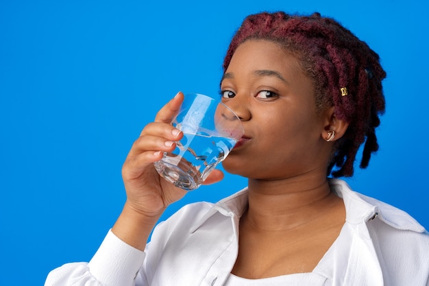 Joven africana bebiendo agua de un vaso contra el fondo azul.
