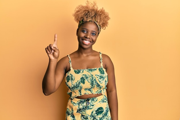 Joven africana con cabello afro vestido de verano que aparece y señala con el dedo número uno mientras sonríe confiada y feliz
