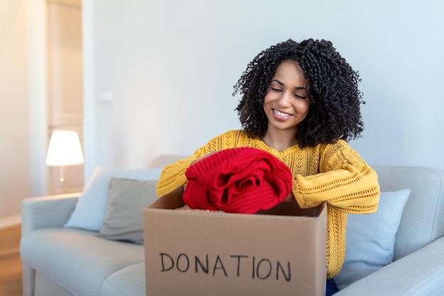 Una joven africana feliz sentada en un sofá pegada a la ropa en una caja de donación en casa, una voluntaria birracial cuidando a una mujer voluntaria poner ropa en un paquete de cartón donar a las personas necesitadas reutilizar el concepto de reciclaje