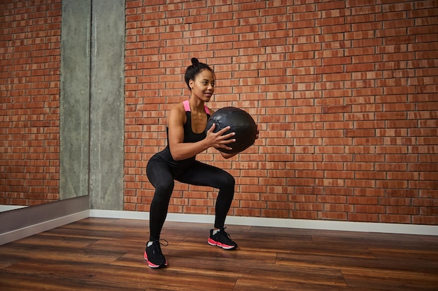 Foto joven africana con hermoso cuerpo musculoso haciendo ejercicios en las nalgas, en cuclillas con una pelota de gimnasia médica mientras entrena en el gimnasio. concepto de estilo de vida activo y saludable