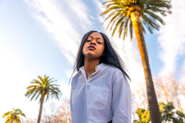 Joven africana posando con ropa blanca en un lugar tropical con palmeras al atardecer disfrutando del verano sonriendo