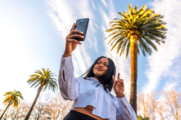 Joven africana posando con ropa blanca en un lugar tropical con palmeras al atardecer tomando una selfie con su teléfono móvil