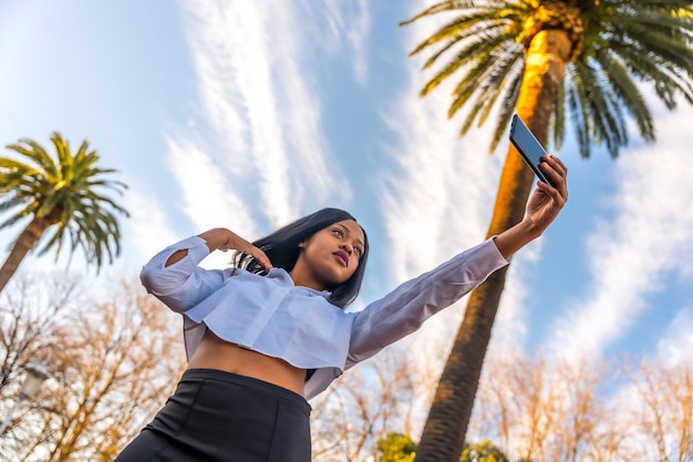 Joven africana posando con ropa blanca en un lugar tropical con palmeras al atardecer tomando una selfie