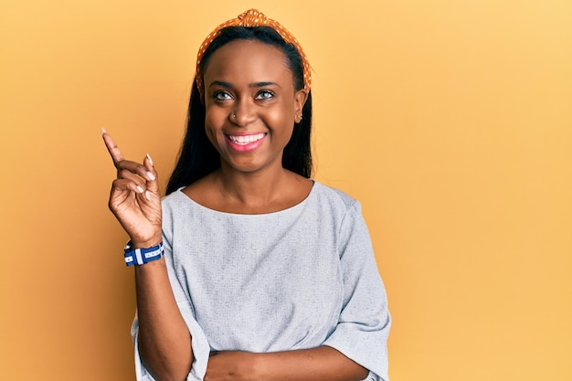 Joven africana con ropa informal de fondo amarillo sonriendo feliz señalando con la mano y el dedo a un lado