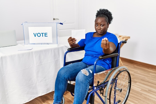 Joven africana sentada en silla de ruedas votando poniendo un sobre en la urna haciendo un gesto italiano con la mano y los dedos expresión segura