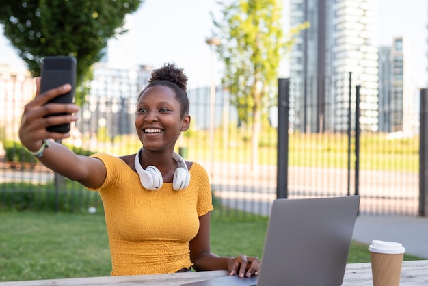 Joven africana tomando una selfie mientras trabaja al aire libre estudiante sentada en una mesa en el parque usando un teléfono inteligente y una computadora portátil múltiples conexiones blogger conectado a internet