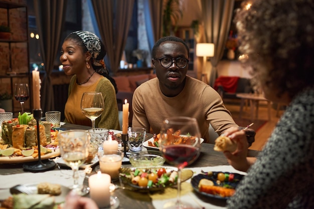 Joven africano en anteojos hablando con sus amigos durante la cena en la mesa