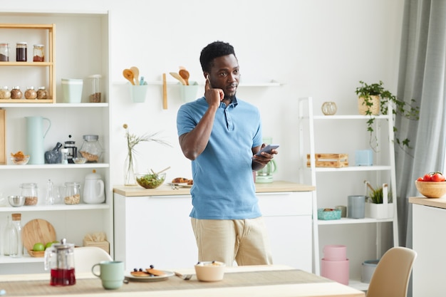 Joven africano con auriculares para hablar por teléfono móvil mientras está de pie en la cocina de casa