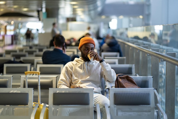 Joven africano bebiendo café y comiendo sándwich mientras espera volar en la terminal del aeropuerto