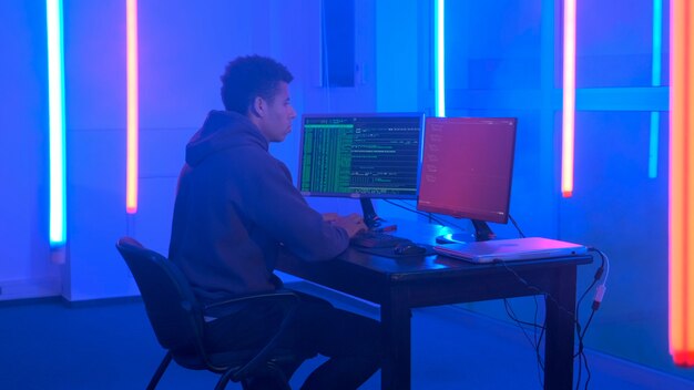 Joven africano escribiendo programas de piratas informáticos en una habitación oscura con luces de neón que trabaja en la computadora con ...