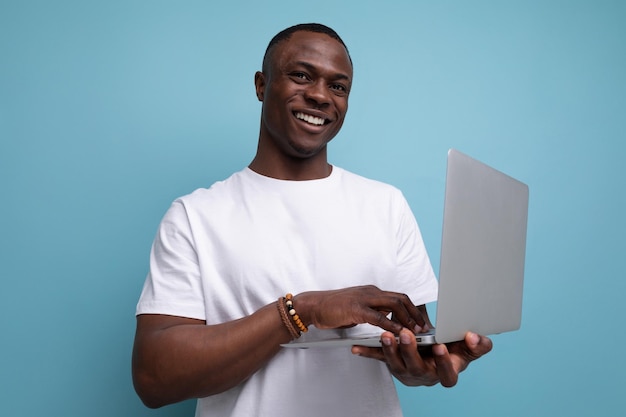 joven africano exitoso en camiseta blanca trabajando remotamente usando portátil en el fondo del estudio con espacio de copia