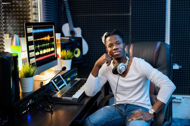 Joven africano exitoso en ropa casual sentado en el lugar de trabajo con monitor de computadora en estudio de grabación de sonido