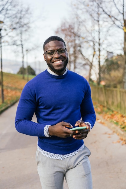 Un joven africano exuda encanto con un suéter azul informal y gafas radiantes de alegría mientras sostiene el teléfono en un retrato bien iluminado del parque