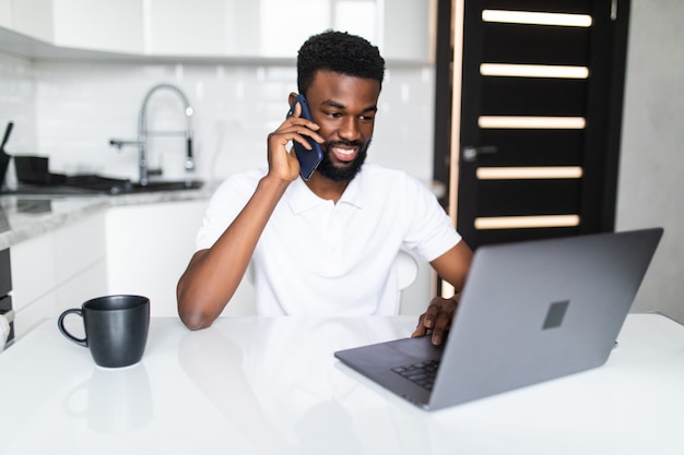 Foto joven africano hablar por teléfono mientras usa la computadora portátil en la cocina