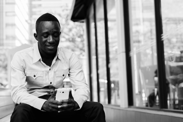 Joven africano negro feliz sonriendo y sentado mientras usa el teléfono móvil fuera de la cafetería con ventanas de vidrio