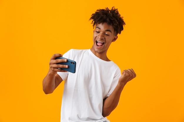 Joven africano posando aislado sobre el espacio amarillo jugar juegos por teléfono hace gesto de ganador.