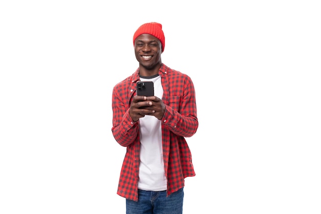 Joven africano positivo con gorra y camisa charlando en un smartphone sobre un fondo blanco aislado