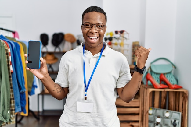 Joven africano que trabaja en una tienda minorista que muestra la pantalla del teléfono inteligente apuntando con el pulgar hacia el lado sonriendo feliz con la boca abierta