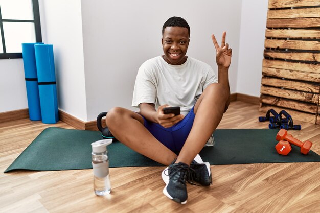 Joven africano sentado en una alfombra de entrenamiento en el gimnasio usando un smartphone sonriendo con cara feliz guiñando un ojo a la cámara haciendo el signo de la victoria con los dedos. número dos.