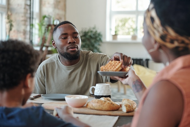 Joven africano sentado junto a la mesa de la cocina mientras toma gofres caseros del plato sostenido por su esposa durante el desayuno familiar en la mañana