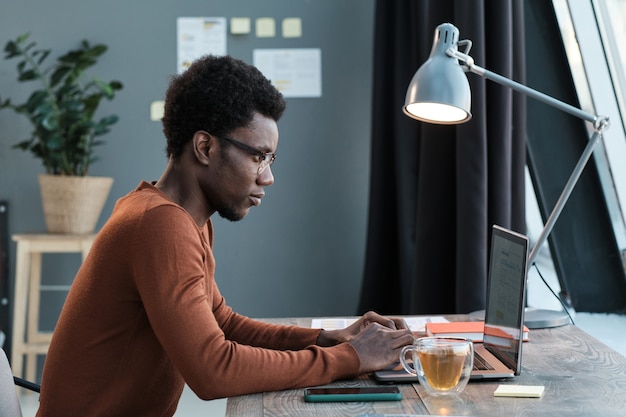 Joven africano sentado en la mesa y escribiendo en la computadora portátil que trabaja en línea en casa