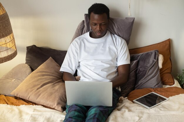 Joven africano sonriente positivo escribiendo en la computadora portátil en casa