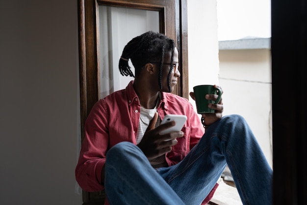 Joven africano sonriente sosteniendo un teléfono inteligente y una taza de té relajándose en silencio junto a la ventana en casa