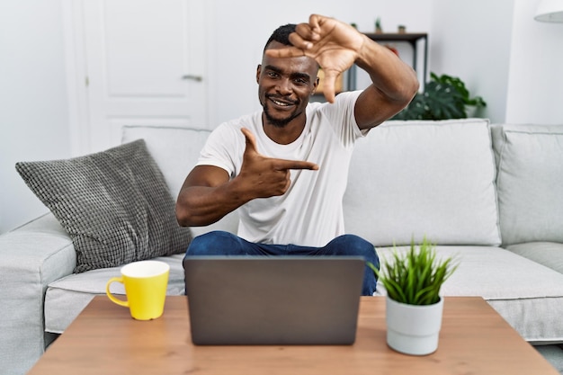 Joven africano usando laptop en casa sonriendo haciendo marco con manos y dedos con cara feliz creatividad y concepto de fotografía