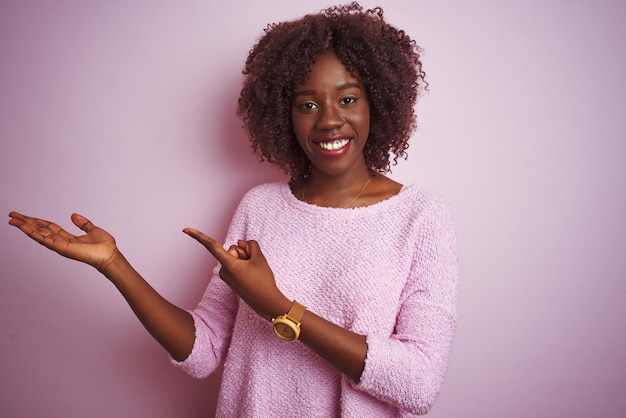 Joven afro africana con suéter de pie sobre un fondo rosado aislado asombrada y sonriendo a la cámara mientras presenta con la mano y señala con el dedo