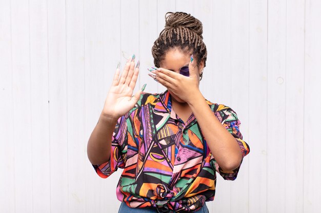 Joven afro cubriéndose la cara con la mano y poniendo la otra mano al frente para detener la cámara, rechazando fotos o imágenes