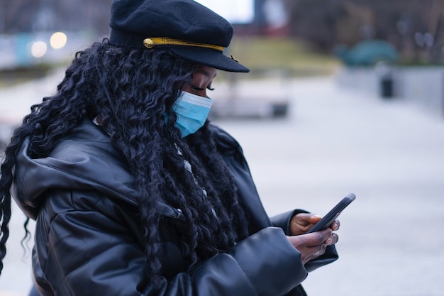 Joven afro con mascarilla médica facial utiliza el teléfono al aire libre