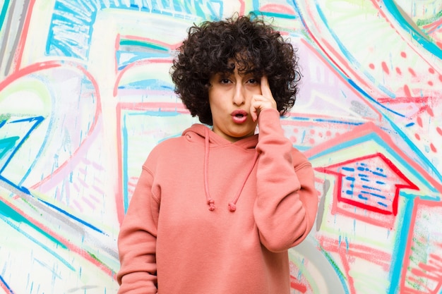 Foto joven afro mirando sorprendida, con la boca abierta, conmocionada, dándose cuenta de un nuevo pensamiento, idea o concepto en la pared de graffiti