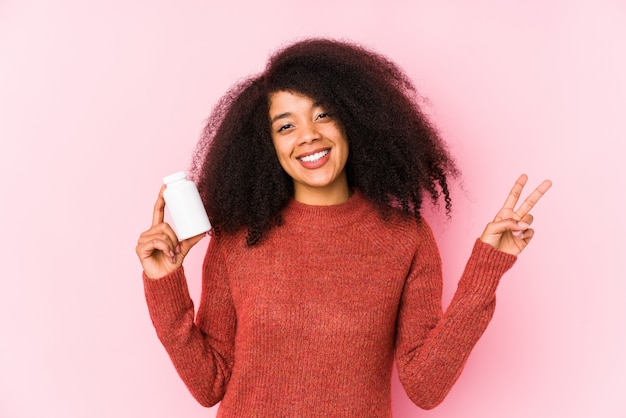 Joven afro mujer sosteniendo un vitaminas aisladas Joven afro mujer sosteniendo un vitaminas alegre y despreocupado mostrando un símbolo de paz con los dedos.