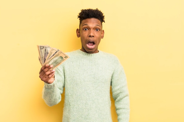 Joven afro negro que parece muy sorprendido o sorprendido por el concepto de billetes de banco en dólares