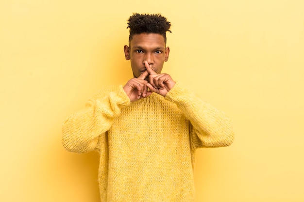 Joven afro negro que parece serio y disgustado con ambos dedos cruzados al frente en señal de rechazo pidiendo silencio