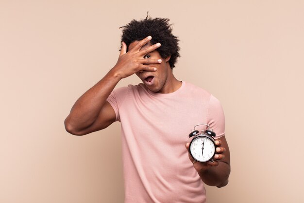 Joven afro negro que parece sorprendido, asustado o aterrorizado, cubriendo la cara con la mano y mirando entre los dedos