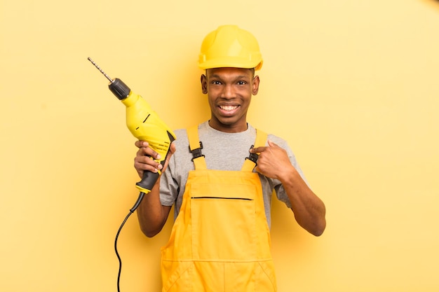 Joven afro negro que se siente feliz y se señala a sí mismo con un trabajador de limpieza emocionado con un taladro