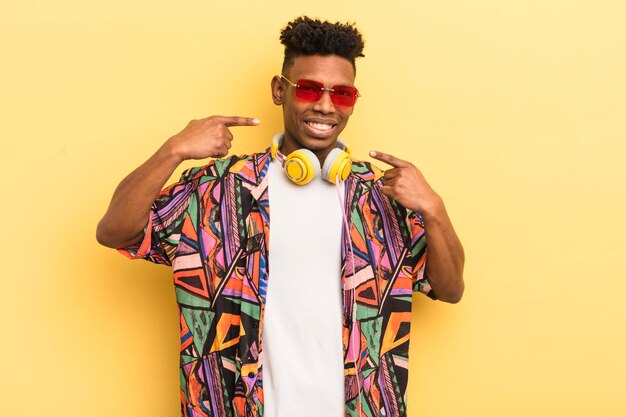 Joven afro negro sonriendo con confianza apuntando a su propia sonrisa amplia con gafas de sol y auriculares