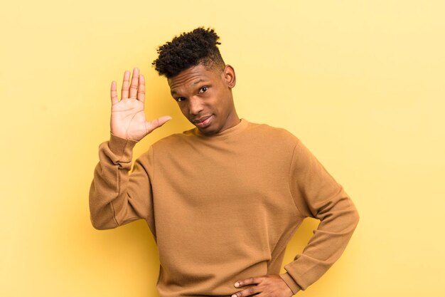 Joven afro negro sonriendo feliz y alegremente agitando la mano dándote la bienvenida y saludándote o despidiéndote